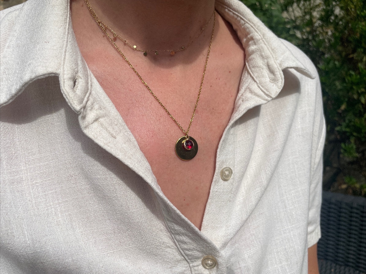 Collier médaille dorée avec pierre de naissance rouge, porté au cou sur une chemise blanche en lin