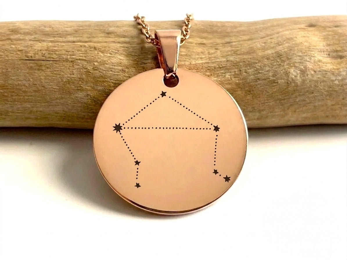 Collier médaille or rose gravée de la constellation de la Balance, posé contre une branche de bois