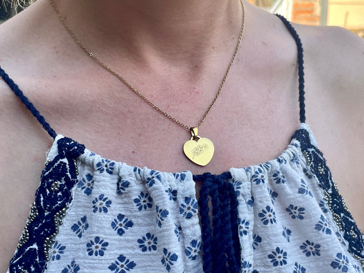 Collier cœur doré gravé d'une fleur, porté au cou sur un haut d'été blanc à fleurs bleues