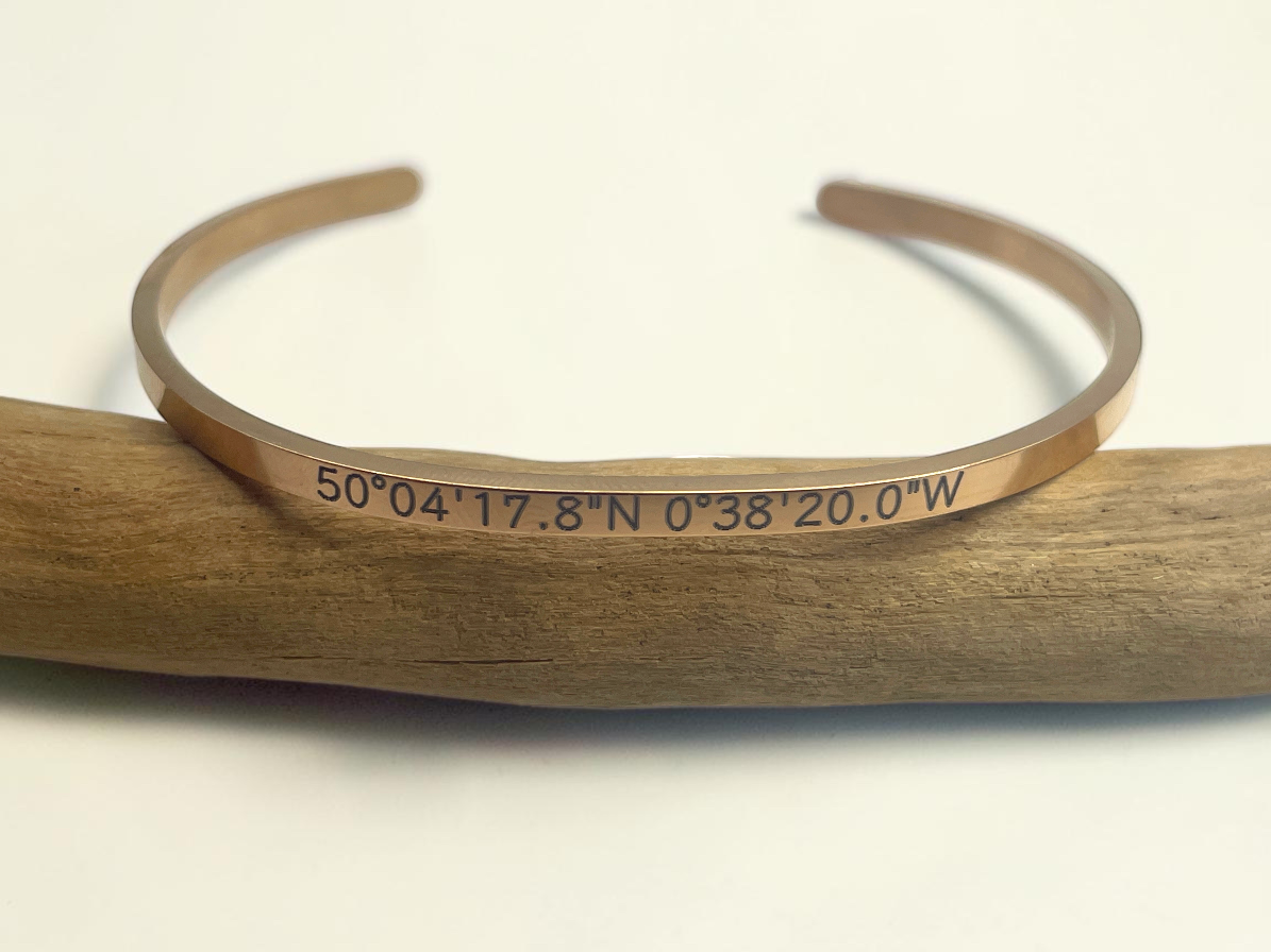 Bracelet jonc doré gravé de coordonnées GPS personnalisées, posé sur une branche de bois