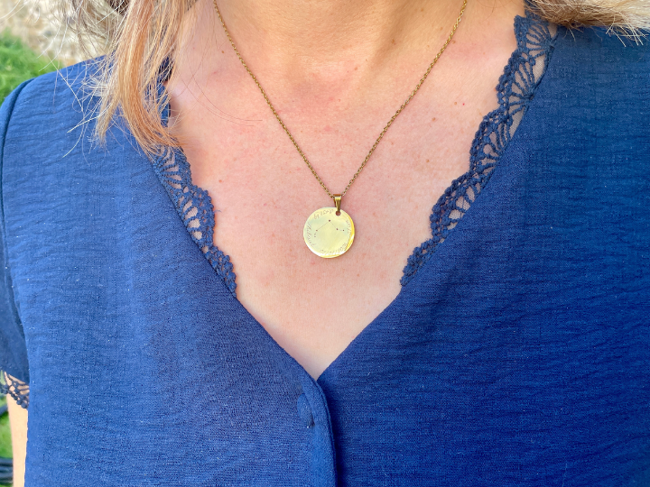 Collier médaille dorée constellation porté au cou, sur un haut bleu en dentelle