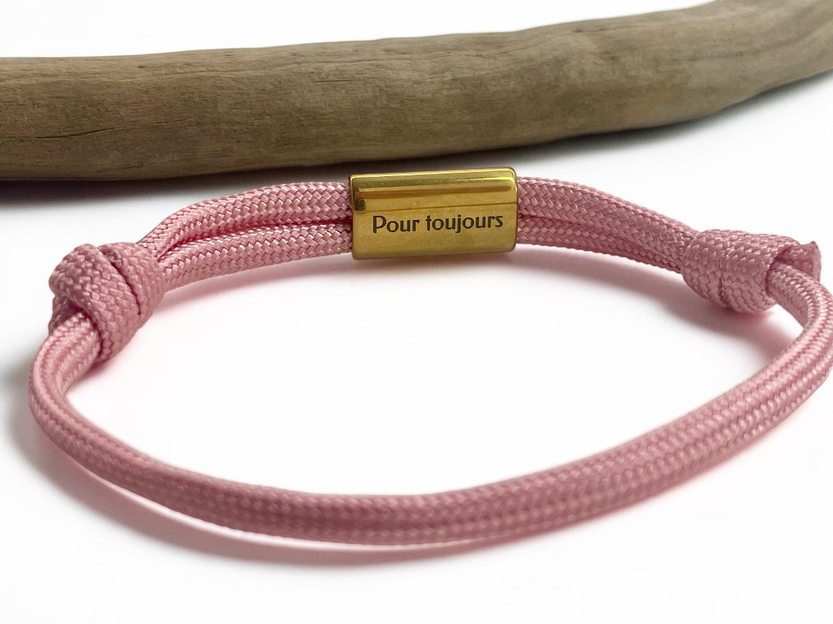 Bracelet cordon rose ajustable à plaque dorée gravée, posé sur une branche de bois