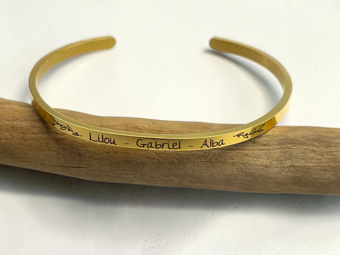 Bracelet jonc doré gravé des prénoms Lilou, Gabriel et Alba, posé sur une branche de bois