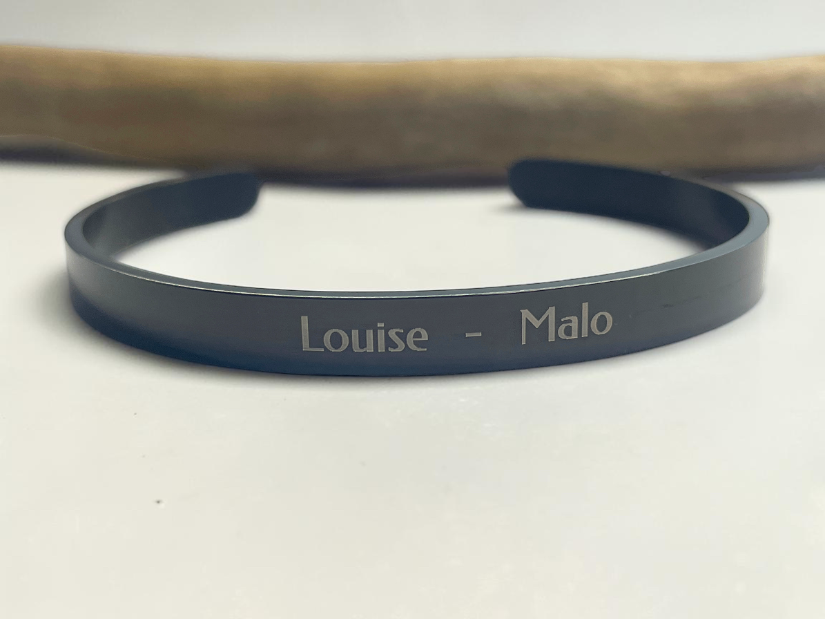 Bracelet jonc noir personnalisé gravé de prénoms, posé sur une branche de bois clair