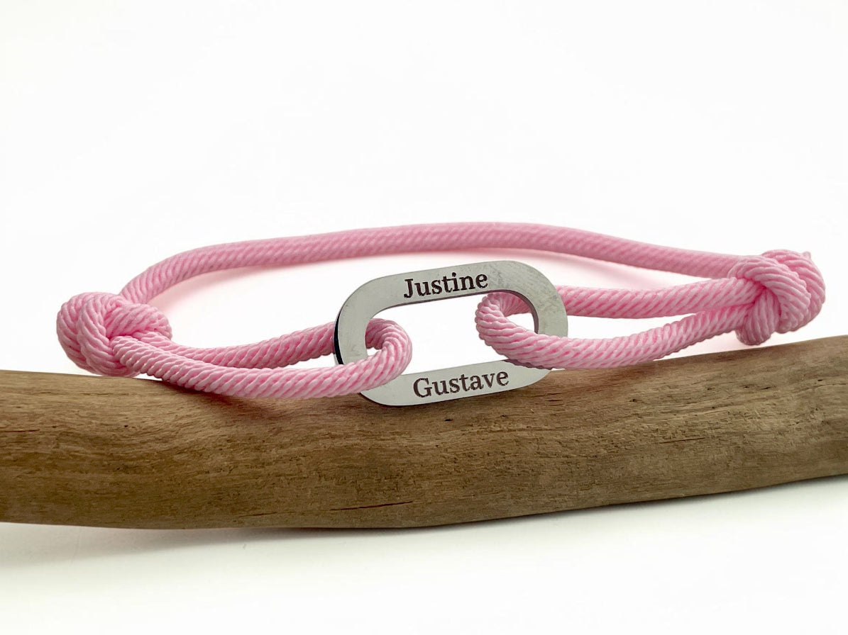 Bracelet cordon rose gravé de prénoms sur son maillon acier, posé sur une branche