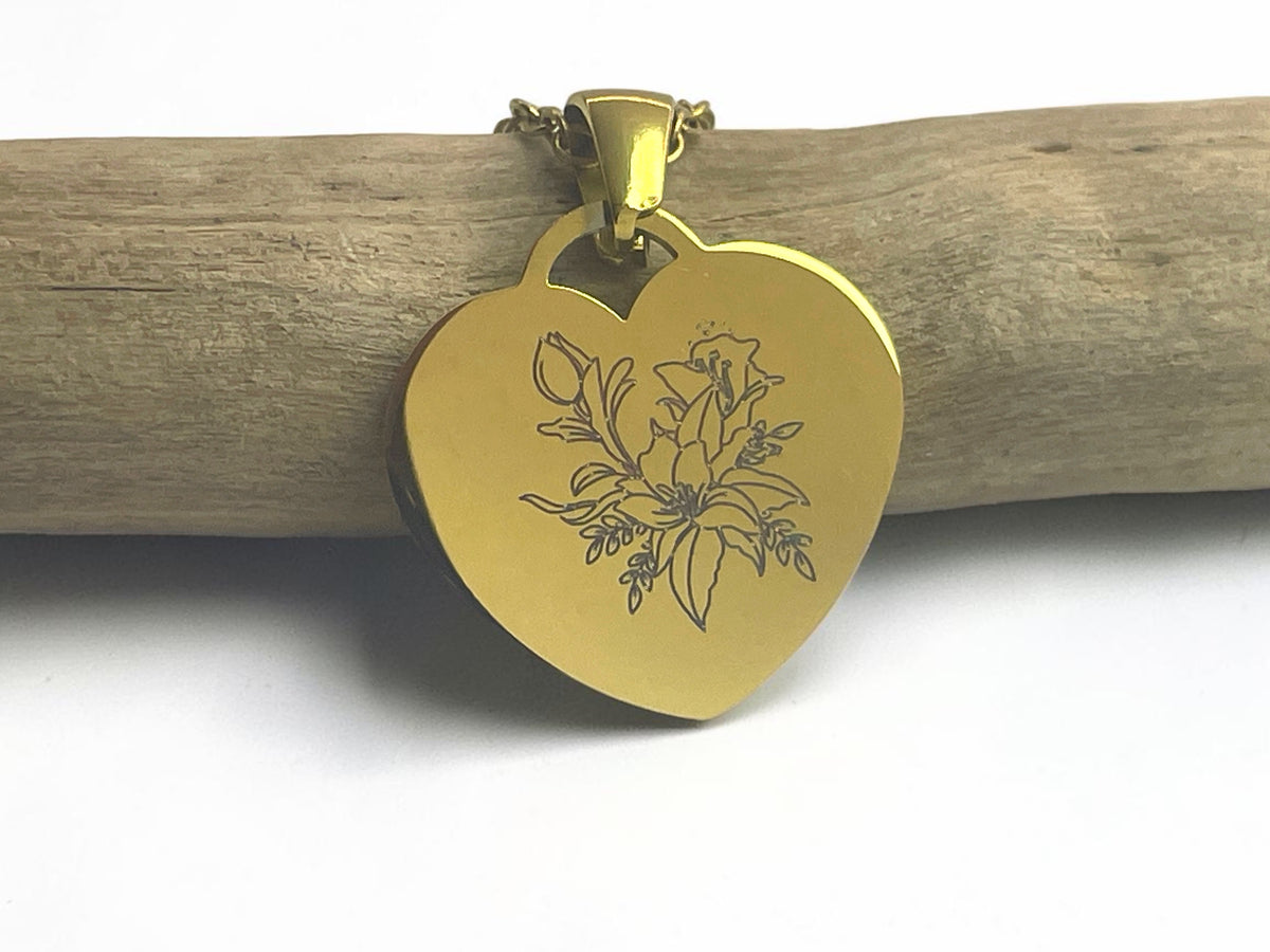 Collier cœur doré gravé d'une fleur au trait fin, suspendu à une branche de bois