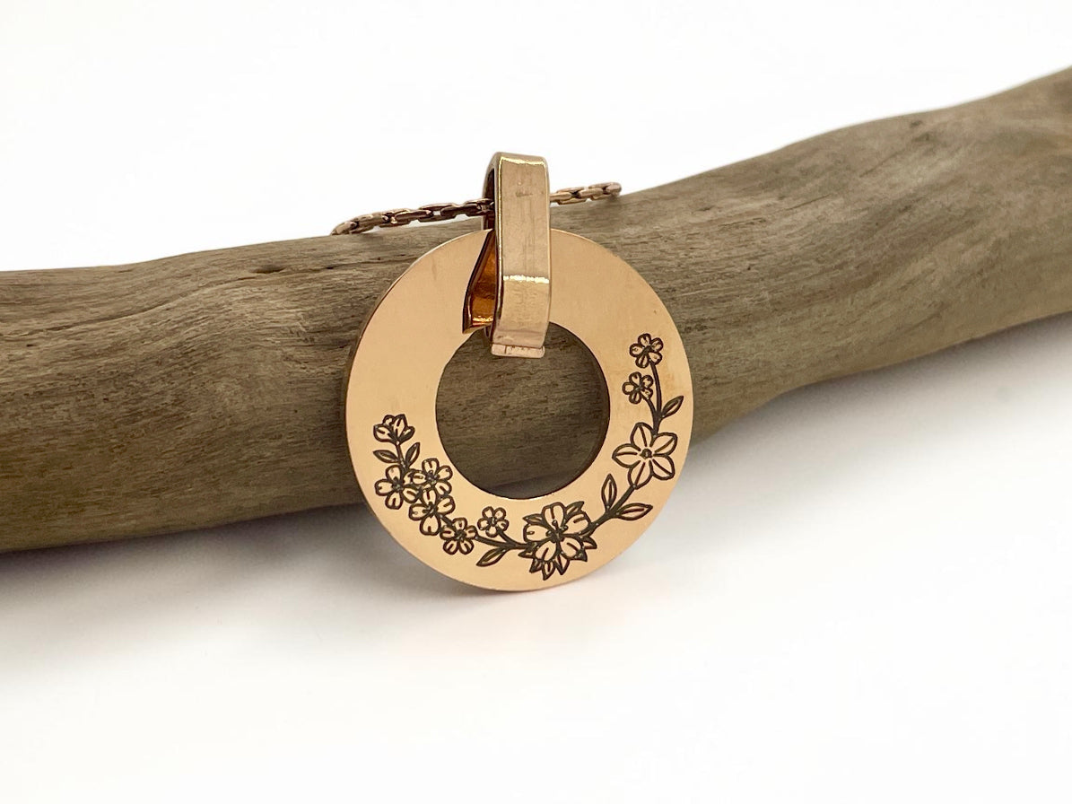 Collier médaille ajourée or rose gravée d'une couronne de fleurs, posé sur une branche