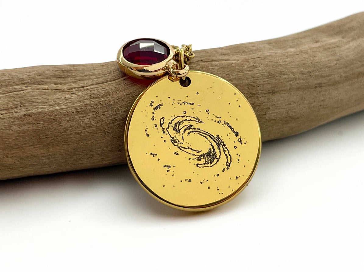 Collier médaille dorée gravée d'une galaxie spirale, avec pierre de naissance rouge