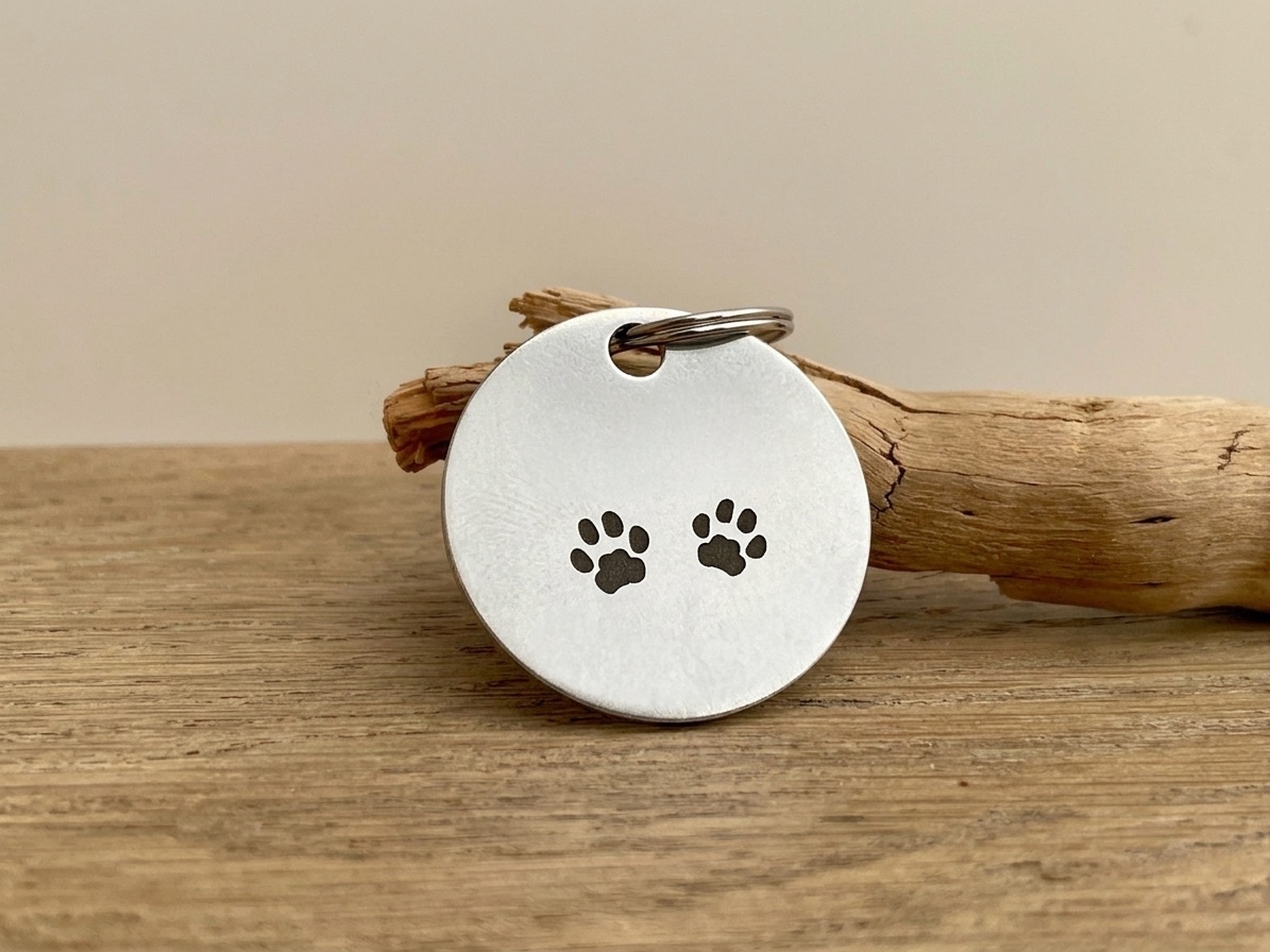 Médaille chien ronde en acier gravée de deux empreintes de pattes, posée contre une branche