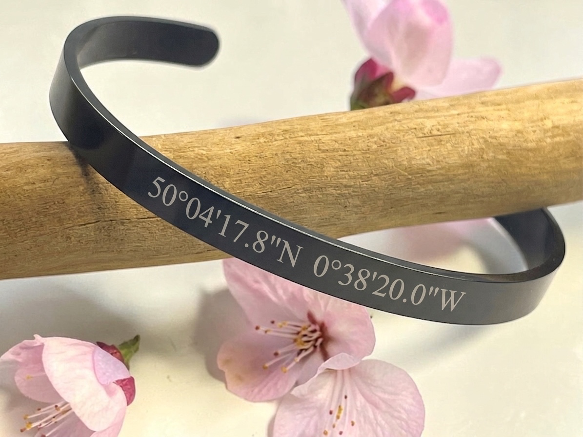 Bracelet jonc noir gravé de coordonnées GPS posé sur une branche, fleurs de cerisier
