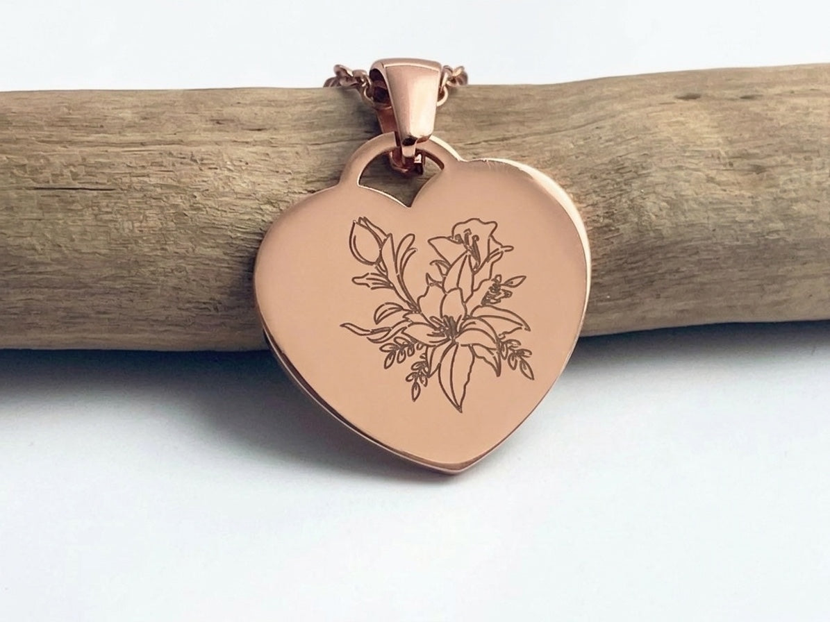 Collier cœur or rose gravé d'une fleur, posé sur une branche de bois clair