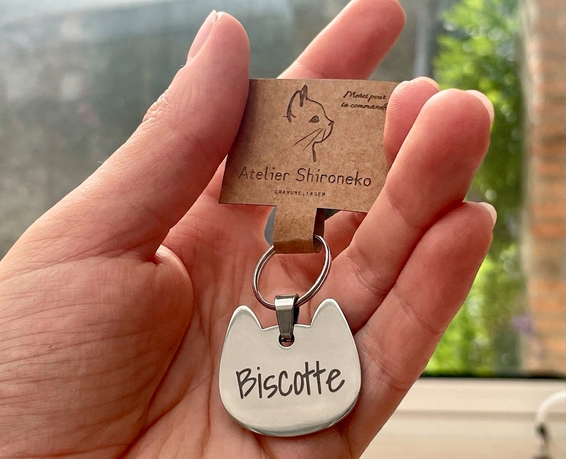 Médaille chat gravée du prénom Biscotte tenue en main avec sa carte Atelier Shironeko
