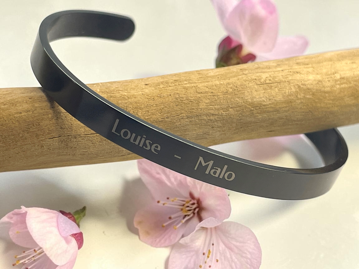 Bracelet jonc noir gravé des prénoms Louise et Malo, posé sur une branche de bois