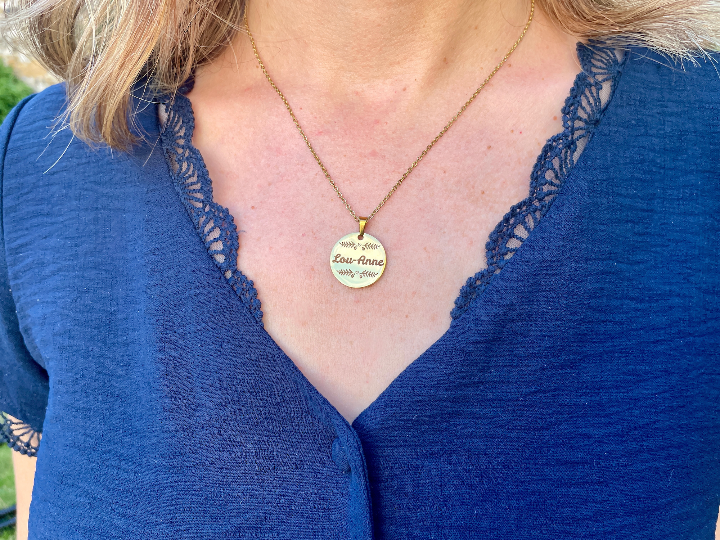 Collier médaille prénom porté au cou, sur un haut bleu en dentelle