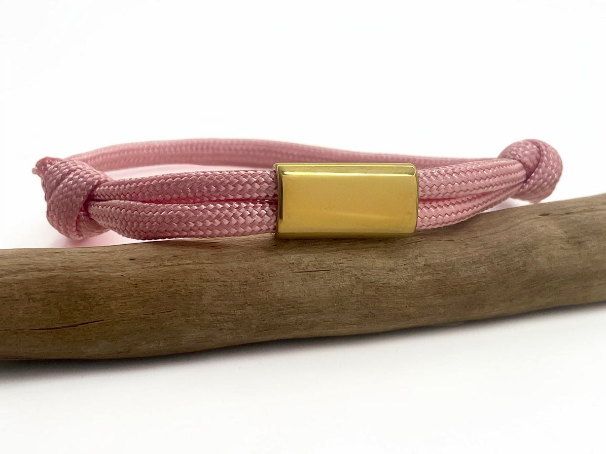 Bracelet cordon rose avec plaque acier dorée à graver, posé sur une branche de bois