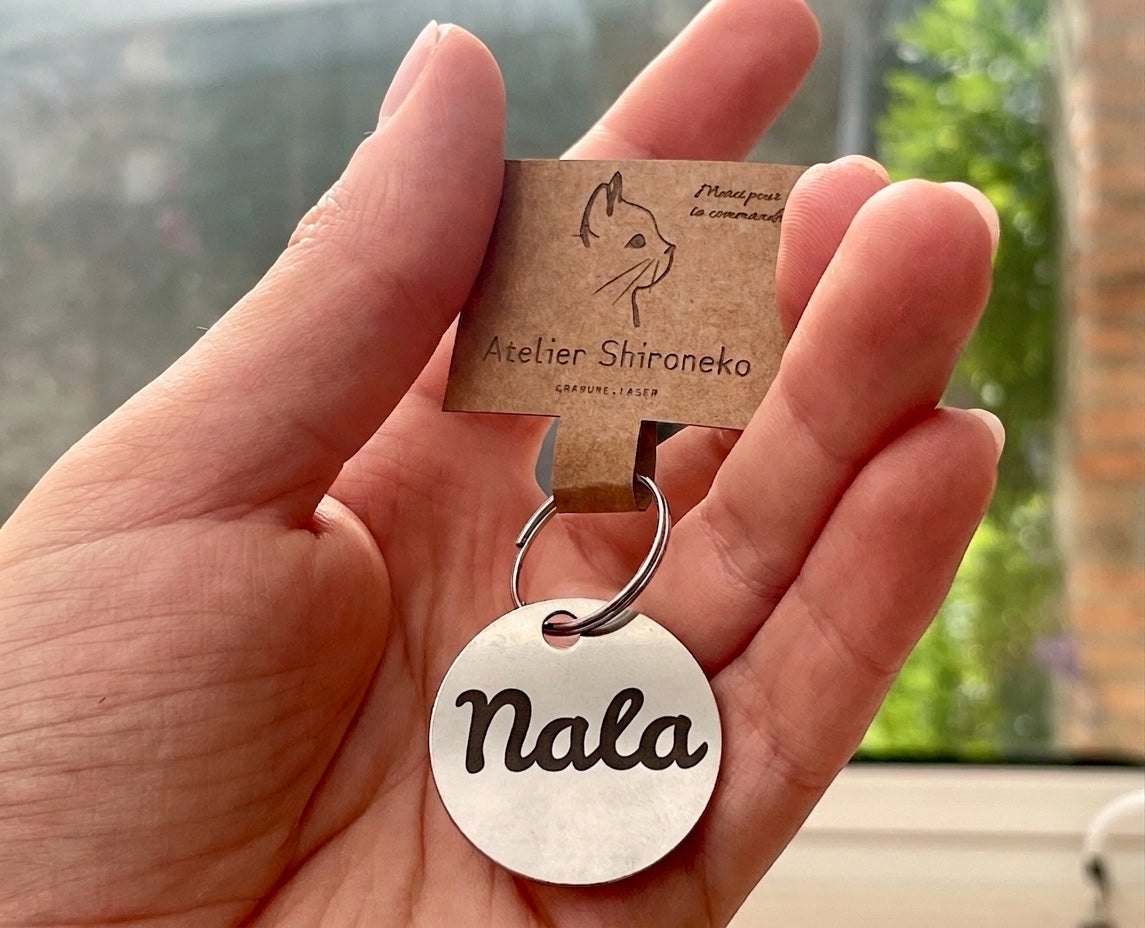 Médaille chien ronde gravée du prénom Nala, tenue en main avec sa carte L'Atelier Shironeko