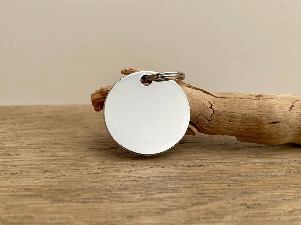 Médaillon d'identité rond en acier inoxydable pour chien, gravé par Atelier Shironeko, posé sur un morceau de bois.